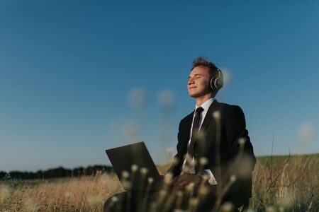 young businessman with headphones listening music in field, sitting on chair, using laptop, closed eyes, outdoorの写真素材