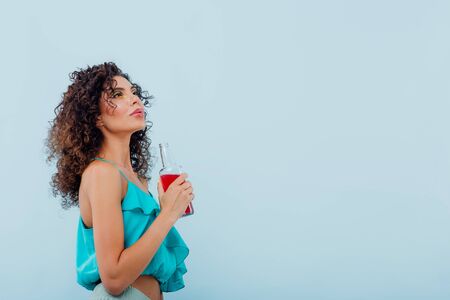 fashionable lady smiles has the drink bottle in her hand, dressed in blue shirt, looks up isolated on blue background, copy spaceの写真素材