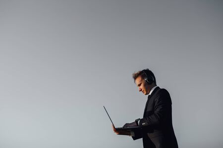 young man using laptop has headphones on his head listening to music, copy space, profile view, sky background, outdoorの写真素材