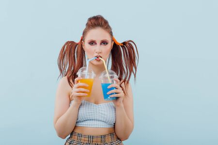 young girl hairstyle and makeup unusual is holding two plastic cups of juice, isolated blue background, positive facial emotions, copy spaceの写真素材