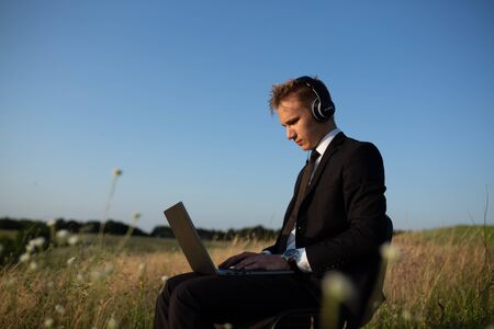 young man with headphones using laptop, in field, sitting on chair, in suit, outdoorの写真素材