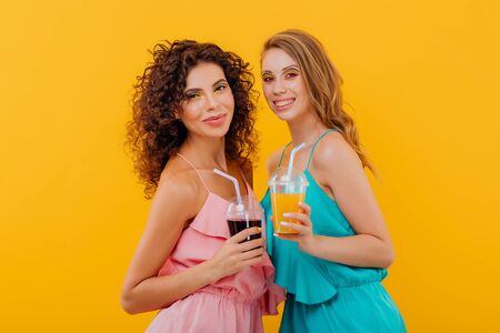fashionable two cheerful girls with curly hair, hold the plastic cup with juice, look at the camera positive facial emotions, isolated on yellow background, copy spaceの写真素材