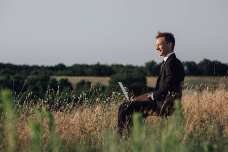 Young happy businessman on meadow,  sitting in the chair, using a laptop, outdoor, profile viewの写真素材