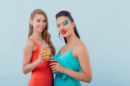two beautiful girls hairstyle, makeup unusual, they drink in hand, looking at the camera, positive facial emotions, isolated on blue background, copy spaceの写真素材