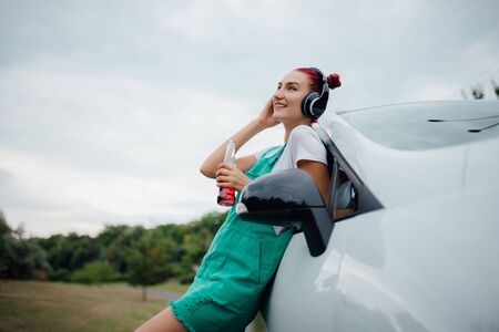 happy young girl with headphones. listen to music, have drink bottle in hand. copy space, next to the car, outdoor, profile viewの写真素材