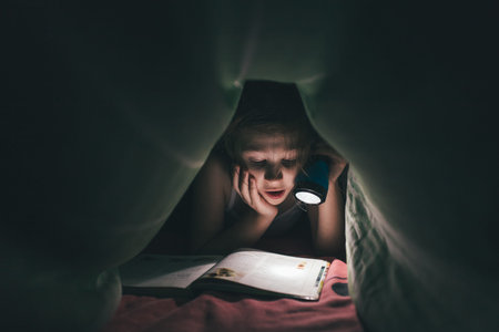 surprised little girl is read a book under a blanket with a flashlight in a dark room at night, in bed, tonの写真素材