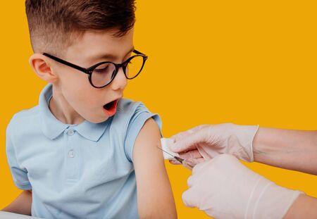 Shocked little boy in glasses waiting for crop nurse to inject vaccine against yellow backgroundの写真素材