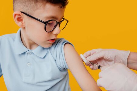 little boy in glasses waiting for crop nurse to inject vaccine against yellow backgroundの写真素材