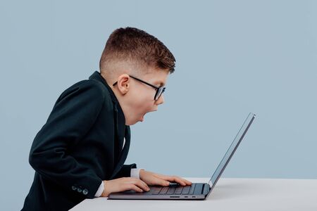 Troubled little schoolboy in suit and glasses and reading data from laptop while working on business project against blue backgroundの写真素材