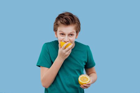 child boy bite from fresh lemon, sour taste, make grimace, facial emotions negative, in green t-shirt isolated on blue background, copy spaceの写真素材