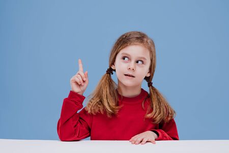 eureka, little girl is sitting at the table with her index finger up. has an idea, dressed in red, isolated on blue background, in studioの写真素材