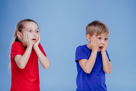 Shocked little kids girl and boy in casual clothes holding hands on faces isolated on blue background, in studioの写真素材