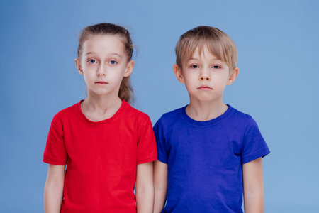 two sad child little girl and boy dressed in T-shirt, isolated on blue background, in studio