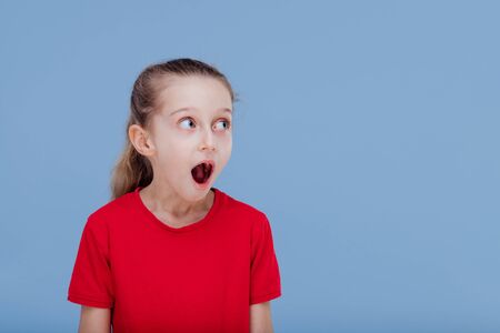 Astonished little girl looking away dressed in red T-shirt, isolated on blue background, in studioの写真素材