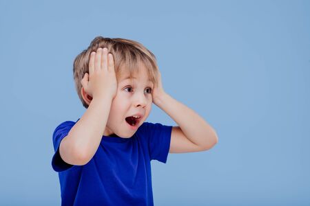 amazed little boy with hands on head looks away dressed in blue T-shirt, isolated on blue background, in studioの写真素材