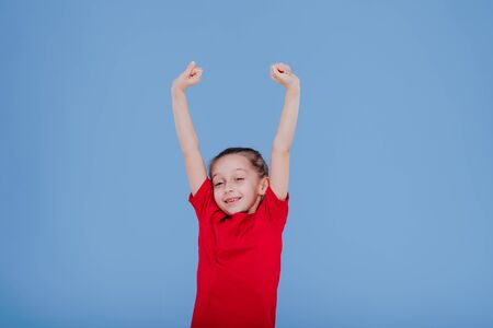 happy little girl in red outfit raising arms and jumping while having fun and celebrating success in studio with blue backgroundの写真素材