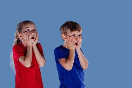 Astonished little girl and boy in casual clothes holding hands on faces and looking away isolated on blue background, in studioの写真素材