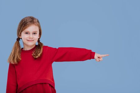 copy space, little girl with smile pointing in left side direction dressed in red, isolated on blue background, in studioの写真素材