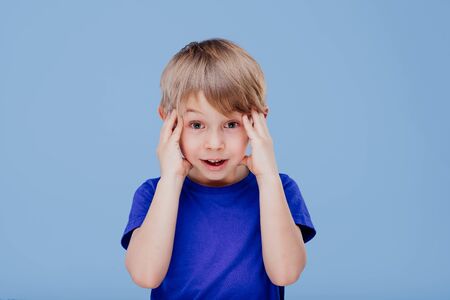 little boy with hands on head looks at camera dressed in blue T-shirt, isolated on blue background, in studioの写真素材