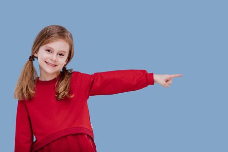 little girl with smile pointing in left side direction dressed in red, isolated on blue background, in studioの写真素材