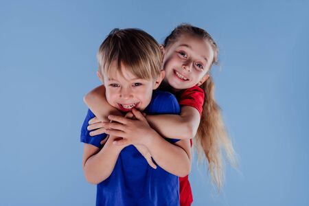 Cute little girl embracing from behind cheerful brother while standing together isolated on blue background, in studioの写真素材
