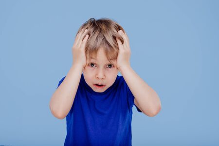 surprised little boy with hands on head looks at camera dressed in blue T-shirt, isolated on blue background, in studioの写真素材