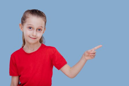Adorable smiling little girl in red t shirt looking at camera and pointing away while standing against blue backgroundの写真素材