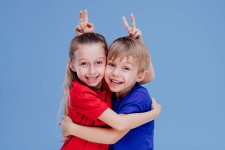 Cheerful cute little girl and boy in bright casual clothes hugging and making horns while looking at camera on blue backgroundの写真素材