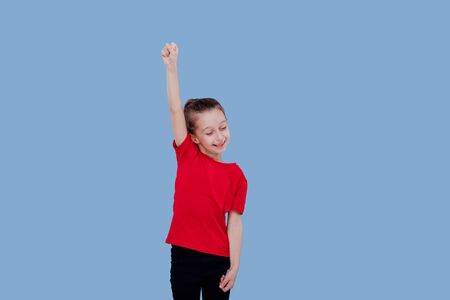 Positive little girl in casual outfit celebrating achievement with raised arm while standing against blue backgroundの写真素材