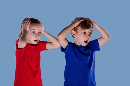 amazed little girl and boy in casual clothes holding head on faces and looking away isolated on blue backgroundの写真素材