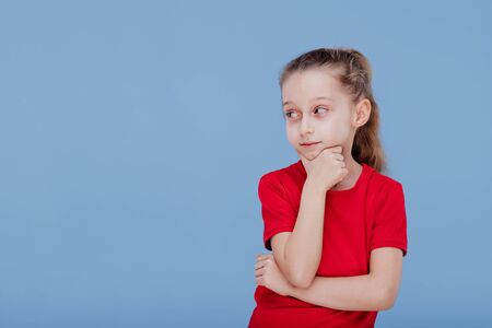 thinking child in red t-shirt touching chin and looking away isolated on blue backgroundの写真素材