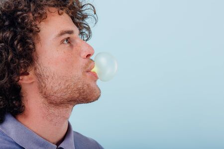 Crop side view of curly man in blue shirt blowing bubble of bubble gum on blue backgroundの写真素材