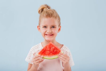 happy caucasian little girl is holding a slice of watermelon. isolated on blue background, copy space, in studio,の写真素材