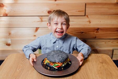 High angle of happy boy smiling and looking at camera while having large burger for lunch in cafeの写真素材
