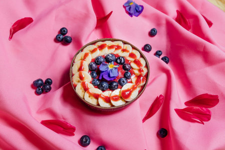 Top view of bowls with sweet fruit dishes placed on pink cloth amidst fresh flowers