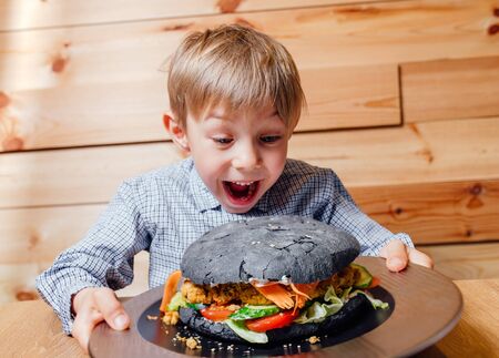 surprised little boy with vegetarian black burger, looking into the camera, wood backgroundの写真素材