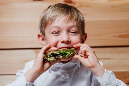 Cheerful boy smiling and biting vegetarian sandwich with vegetables while having lunch against wooden wallの写真素材