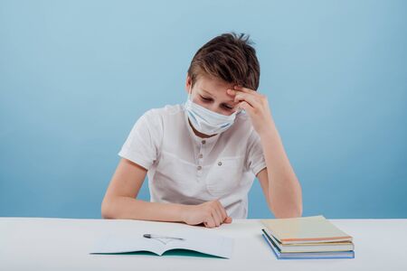 tired schoolboy with medical mask your hands on your head, sitting at the table, isolated on blue backgroundの写真素材