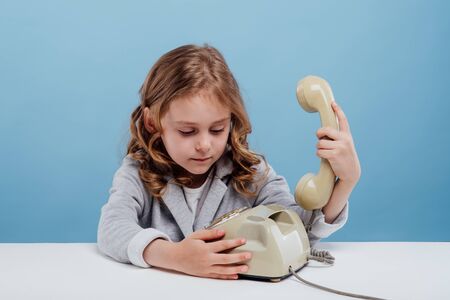 confused little girl with old telephone, sitting at the table, blue backgroundの写真素材