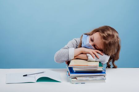 Exhausted preteen pupil in medical face mask resting on stack of books and copybooks while sitting at white tableの写真素材
