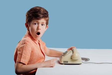 Side view of amazed little male child in orange shirt looking at camera, and showing old phone put at white table on blueの写真素材