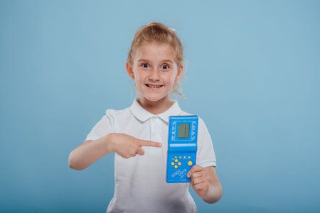 Happy surprised preteen girl holding, and point finger on blue retro electronic portable game...