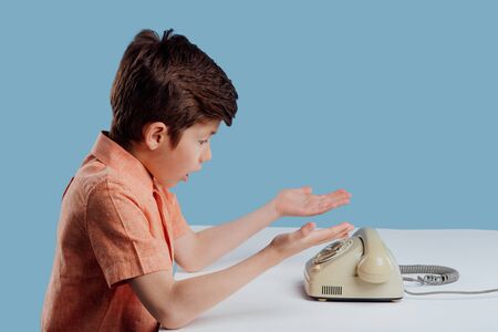 Side view of stylish amazed little child in orange shirt, sitting with palms up and looking at old fashioned telephone put on table on blue backgroundの写真素材