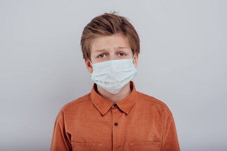 Unhappy exhausted teenager in orange shirt and protective mask looking at camera while standing alone against grayの写真素材