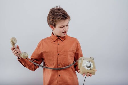 Dissatisfied surprised preteen boy, in orange formal shirt holding and looking at retro disk phone with aversion and misunderstand against grayの写真素材