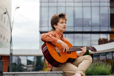 child plays guitar in the park, bottom view, dressed in a shirt, city background dressed in a shirt, building in background, profile viewの写真素材