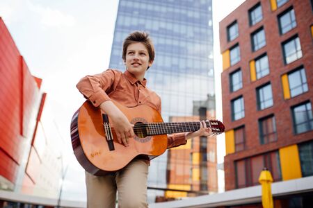Handsome preteen plays guitar in the park, bottom view, dressed in a shirt, city backgroundの写真素材