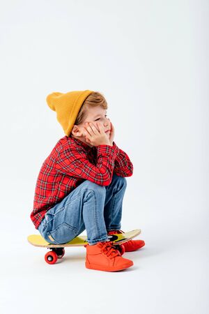 tired child is sitting on a skateboard, dressed in red shirt with squares and jeans, isolated on white background, studio shotの写真素材