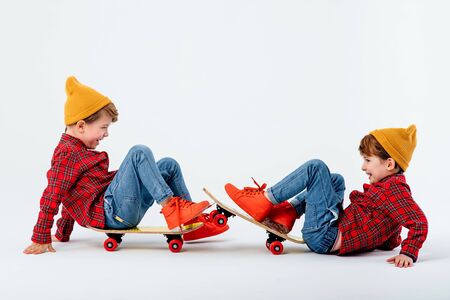 Happy children are sitting on skateboards and playing, dressed in red shirt with squares and jeans, isolated on white background, studio shotの写真素材