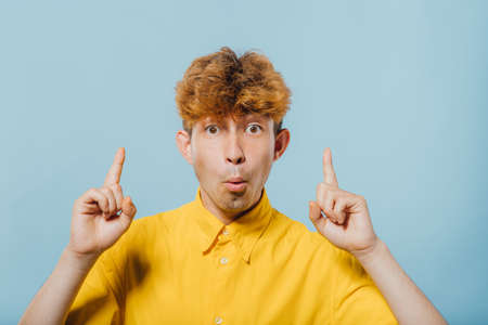 surprised boy pointing fingers up, at copy space, isolated over blue background, teenager with fashionable hairstyle, handsome guy in yellow shirt, young boy, nice teenager,の写真素材